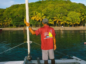 A crew member stands on the deck of a sailing boat, overlooking a beautiful beach scene with guests enjoying the water — a perfect day for sailing adventures.