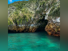 A stunning view of a rocky coastline with lush greenery and a cave entrance, set against the backdrop of crystal-clear turquoise waters — a perfect destination for exploration and adventure.