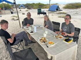 A cheerful group enjoys a meal together at an outdoor dining setup on the beach, perfect for family bonding and adventure trips.