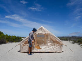 A person is seen interacting with a tent on a sandy beach, surrounded by natural vegetation under a clear blue sky.