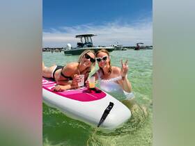 Two friends enjoy a fun day on the water, sipping cocktails while paddleboarding in clear waters — perfect for summer celebrations and beach outings.