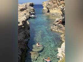 Two individuals enjoy swimming in crystal-clear waters near a rocky coastline, with a boat anchored nearby — a perfect scene for summer adventures and exploration.