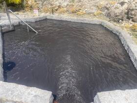 An unclear image of a dark water pool, possibly a natural hot spring or mud bath, surrounded by rocky terrain.