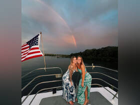 Two friends pose on the deck of a boat with a stunning rainbow in the background, capturing a perfect moment of joy and celebration on the water.