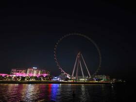 A nighttime view of a cityscape featuring a large Ferris wheel illuminated with colorful lights, reflecting on the water.