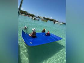 A family enjoys a sunny day on a floating mat in clear turquoise waters, surrounded by boats — perfect for summer fun and relaxation.