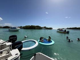A lively scene at a sandbar with friends enjoying the sun, swimming, and relaxing on floating mats while boats cruise nearby — the perfect day on the water.