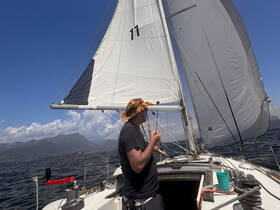 A man adjusts the sails on a sailboat, enjoying a beautiful day on the water with stunning views of the mountains in the background.