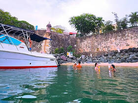 A lively group enjoys a refreshing swim near a boat, surrounded by scenic rocky shores — perfect for a fun day at the beach.