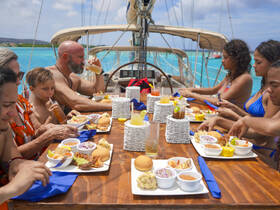 A lively group enjoys a delicious meal aboard a sailboat, surrounded by stunning turquoise waters — perfect for family outings or group sailing adventures.
