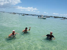 A lively group enjoys a refreshing swim in clear waters, surrounded by boats — perfect for a fun day at the beach.
