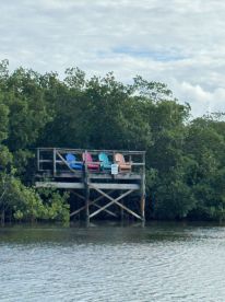 A view of a wooden platform with colorful chairs overlooking the water, surrounded by mangroves. The scene captures a tranquil spot for relaxation, though details are not clearly visible.