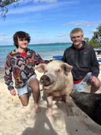 Two young men interact with a friendly pig on the beach, showcasing a unique wildlife experience in the Bahamas.