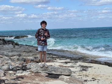 A young man stands on rocky terrain by the ocean, enjoying the scenic view of turquoise waters and distant islands — a perfect snapshot of a beach adventure.