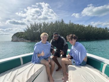 A group of young men enjoy a fun moment on a boat, giving thumbs up against a scenic backdrop of turquoise waters and lush greenery.