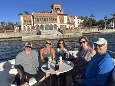 A cheerful group of friends enjoys a day on the water, celebrating with drinks and laughter against the backdrop of a stunning waterfront mansion.