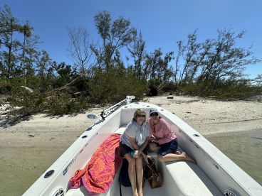 Two friends enjoy a relaxing moment aboard their boat near a sandy beach, surrounded by lush greenery — a perfect getaway for sun and fun.