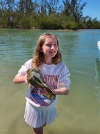 A young girl joyfully holds a large oyster while standing in shallow water, showcasing a memorable wildlife encounter during a fun day on the water.