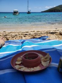 A relaxing beach scene featuring snacks on a blue mat, with boats anchored in the background and a swimmer enjoying the clear waters — perfect for a day of fun in the sun.