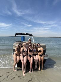 A fun group of friends poses on the beach in front of a pontoon boat, enjoying a sunny day on the water — perfect for summer parties and beach outings.
