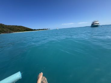 A serene view of turquoise waters with a foot dangling in the water, showcasing a beautiful beach and island backdrop — perfect for relaxing moments on a boat trip.