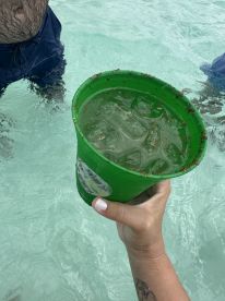 A hand holds a vibrant green cup filled with a refreshing drink while standing in shallow water, perfect for enjoying cocktails during a day on the water.