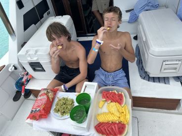 Two boys enjoy a delicious spread of snacks, including guacamole, tortilla chips, and fresh fruit, aboard a boat — perfect for a fun day on the water.