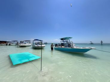 Guests enjoy a sunny day at a sandbar, with multiple boats anchored nearby and a floating mat for relaxation — a perfect setting for summer fun on the water.