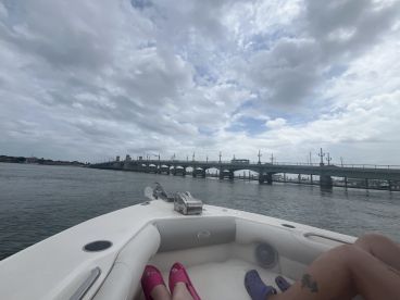 A scenic view from a boat showcasing a bridge in the distance under a cloudy sky, with feet visible on the boat's edge, capturing a moment of relaxation on the water.