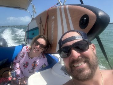 A cheerful couple enjoys a sunny day on a boat, with a paddleboard in the background, capturing the essence of fun on the water.