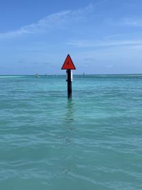 A navigational marker in shallow turquoise waters, indicating a channel or area for boating. The scene captures a serene coastal environment.