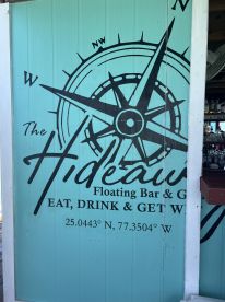 A close-up view of a sign for 'The Hideaway' Floating Bar & Grill, featuring a compass design and coordinates, ideal for visitors looking for a unique dining experience on the water.
