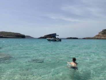 A child swims in the crystal-clear waters of Malta, with a boat anchored nearby, showcasing the perfect beach day and family fun.