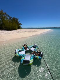Two friends relax on a colorful floating mat in crystal-clear waters, enjoying a sunny day at the beach — a perfect setting for summer fun and relaxation.
