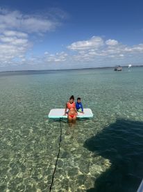 A mother and her son enjoy a sunny day on a floating mat in shallow, clear waters — a perfect family moment for summer fun.