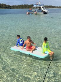 Three kids enjoy a sunny day on a floating mat in clear waters, with a party boat and other guests in the background — perfect for family fun and water activities.