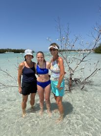 A joyful group of three women, including a young girl, poses in shallow water surrounded by beautiful scenery — a perfect moment of family fun at the beach.