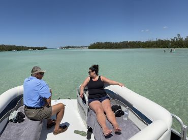 A couple relaxes on a pontoon boat in clear waters, enjoying a sunny day while others wade nearby — a perfect sandbar stop for fun and relaxation.