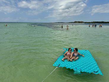 Two friends relax on a floating mat in shallow waters, surrounded by others enjoying a sunny beach day — a perfect scene for summer fun and relaxation.