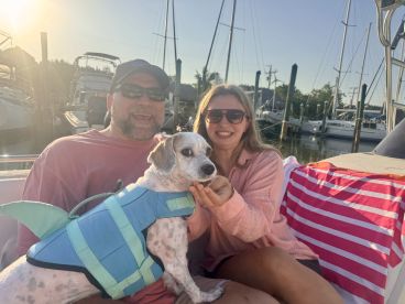 A joyful moment on a boat as a couple poses with their dog, showcasing a fun day on the water. The dog is wearing a life jacket, adding to the playful atmosphere.