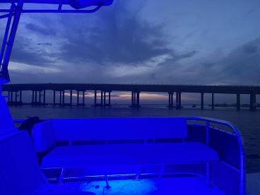 A serene view from the interior of a boat, showcasing comfortable seating and ambient blue lighting against a backdrop of a bridge at sunset.