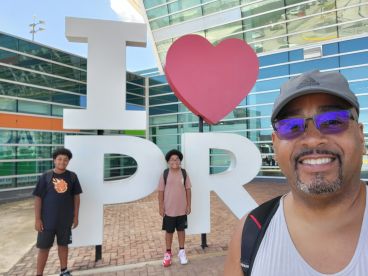A joyful family poses in front of the iconic 'I love PR' sign in Puerto Rico, capturing a memorable moment during their trip.