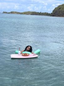 A woman relaxes on a floating mat in clear waters, enjoying a meal and drinks — a perfect scene for a leisurely day on the water.