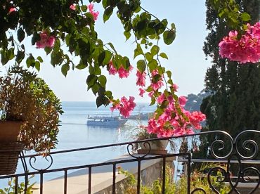 A picturesque view featuring a boat gliding across calm waters, framed by vibrant flowers and lush greenery, capturing the serene beauty of the coastline.