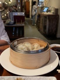 A dim sum dish featuring dumplings in a bamboo steamer, with a restaurant setting in the background.