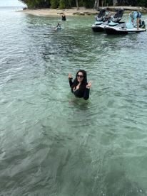 A joyful woman poses for the camera while swimming in clear waters, with friends enjoying jet skis in the background — a perfect day of fun and adventure at the beach.