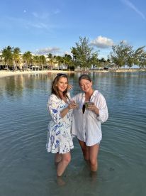 Two friends enjoy cocktails while standing in shallow water, celebrating their beach getaway — a perfect moment for relaxation and fun in the sun.