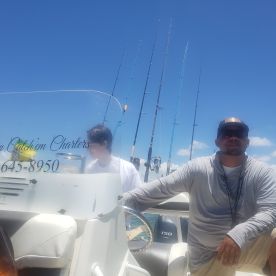 Two individuals aboard a fishing charter boat, ready for a day of fishing under the bright blue sky. The boat's contact information is visible, making it easy to book a fishing adventure.