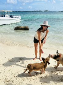 A woman joyfully feeds pigs on a sandy beach, showcasing a unique wildlife interaction in a tropical setting.