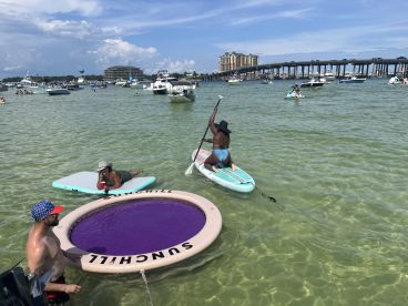 A lively scene at the beach with friends enjoying paddleboarding and relaxing on floating mats in clear waters — perfect for summer fun and water activities.